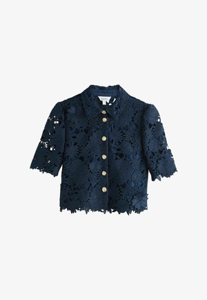 Femme aux longs cheveux blonds portant une robe midi en dentelle florale bleu marine et des sandales à talons noires, marchant devant un fond clair uni.