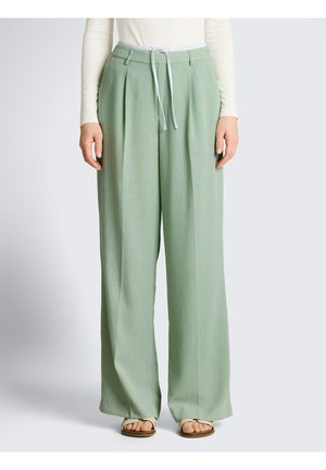 Modelka w luźnych spodniach w kolorze szałwii z przodu plisami, białą ściągaczem w pasie, stylizowana z białym długim rękawem i beżowymi sandałami.