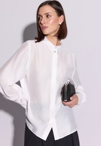 Biała, przezroczysta koszula zapinana na guziki z kołnierzykiem i ozdobnymi guzikami. W jednej ręce trzyma czarną kopertówkę z metalowym zapięciem.