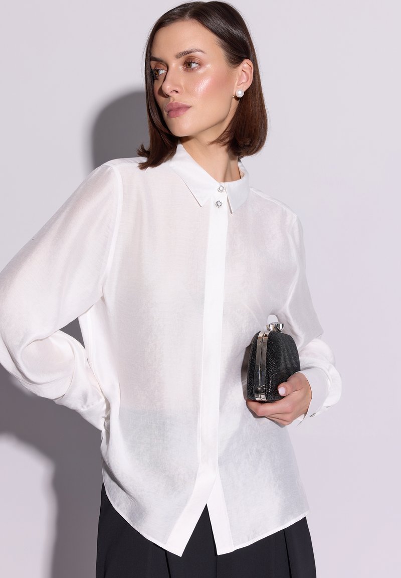 Biała, przezroczysta koszula zapinana na guziki z kołnierzykiem i ozdobnymi guzikami. W jednej ręce trzyma czarną kopertówkę z metalowym zapięciem.