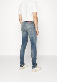 Jeans de mezclilla azul con un ajuste slim, que cuentan con bolsillos traseros y un parche de cuero con el logo. Combinados con una camiseta blanca y zapatillas casuales.