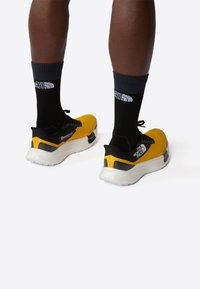 Zapatillas deportivas amarillas con detalles en negro, parte superior de malla, suela acolchada y detalle del logo. Combinadas con calcetines negros que tienen un logo blanco.