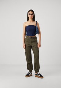 Trumpu mėlyno vamzdžio viršus, alyvuogių žalios spalvos krovinių kelnės su šoninėmis kišenėmis, juodi sportbačiai su baltais dryžiais, mėlynas raštuotas rankinukas ir saulės akiniai.