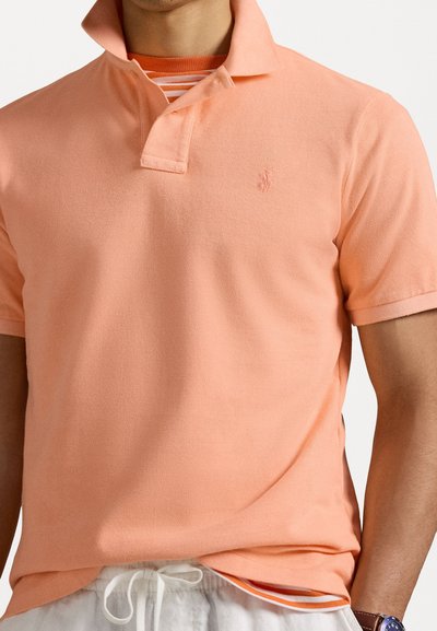 Polo a maniche corte color pesca chiaro con trama testurizzata, colletto e logo ricamato. Indossato sopra una maglietta a righe con pantaloni corti bianchi.