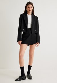 Blazer nero corto con due bottoni, abbinato a pantaloni corti neri a vita alta. Le scarpe sono mocassini neri, completati da calze nere fino al ginocchio.