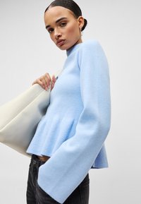 Ανοιχτό μπλε πλεκτό πουλόβερ με ψηλό λαιμό, φαρδιές μακριές μανσέτες και μπάσκα στη μέση. Συνδυασμένο με κρεμ τσάντα.