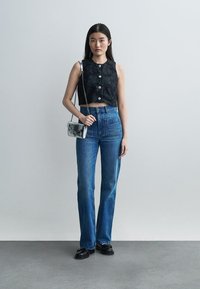 Nainen seisoo yksinkertaisen seinän vieressä, yllään musta hihaton lyhyt paita, jossa kukkakirjailulla koristellut napit, korkeavyötäröiset siniset farkut, mustat kengät ja hopeanvärinen käsilaukku.