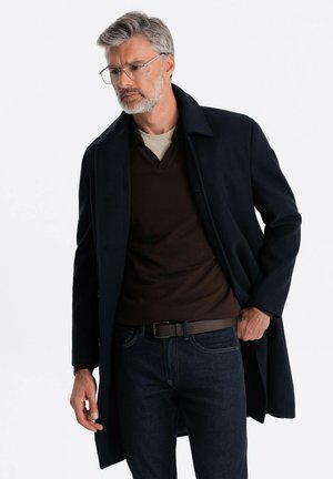 Mørkeblå frakke med et struktureret design, lagdelt over en brun sweater. Har en klassisk krave og er parret med mørke jeans.