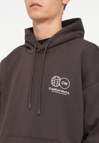 Brun hætte sweatshirt lavet af blødt stof, med en frontlomme, snøre i hætten og et hvidt logo med "Creative Works."