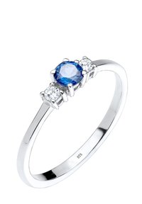 Silverring med en central blå ädelsten flankeras av två mindre vita stenar, polerad yta, enkel banddesign.