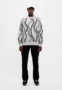 Maglione grigio con motivo di testo ondulato nero, colletto rotondo e polsini a costine, abbinato a pantaloni neri e sneakers nere. Cappellino bianco indossato.