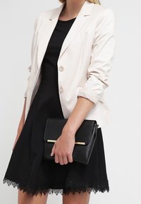 Blazer rosa chiaro con due bottoni, indossato sopra un abito nero. Borsa nera con un accento dorato tenuta in mano. Orlo dell'abito rifinito in pizzo.