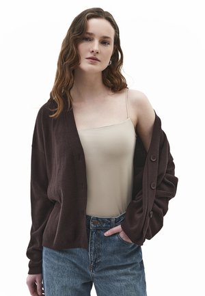 Femme aux cheveux bruns ondulés portant un débardeur beige, un cardigan marron foncé glissant d'une épaule, et un jean bleu, une main dans la poche.