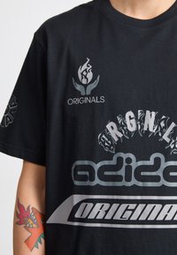 Melnā kokvilnas T-kreklā ir liela pelēka "adidas" logo, teksts "ORIGINALS" un liesmas grafika. Īsas piedurknes ar rakstainiem detaļām sānos.