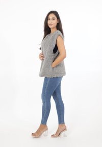 Szary kamizelka z futra syntetycznego o bezrękawym kroju, z bocznymi kieszeniami i miękką teksturą, połączona z niebieskimi dżinsami typu skinny oraz przezroczystymi sandałami na obcasie.