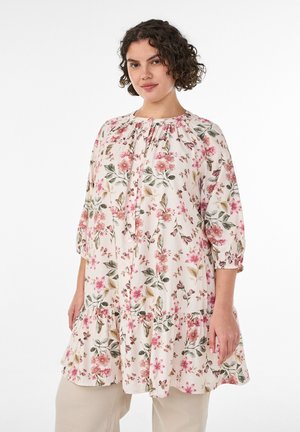 Kvinde med kort krøllet hår iført en løs tunika med blomster i pink og grøn og beige bukser, stående mod en hvid baggrund.