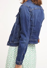 Kvinna som bär en blå jeansjacka över en vit klänning med grönt kaktusmönster, sedd från sidan och uppifrån midjan.