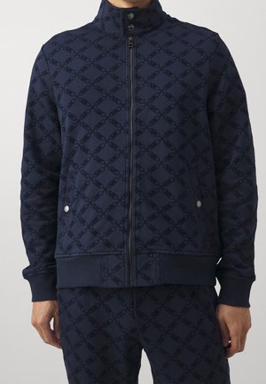 Homme portant une veste zippée bleu marine et un pantalon assorti avec un motif subtil de chaînes entrelacées et des poches à boutons-pression.