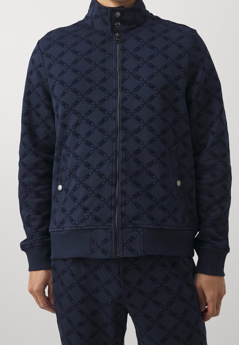 Homme portant une veste zippée bleu marine et un pantalon assorti avec un motif subtil de chaînes entrelacées et des poches à boutons-pression.