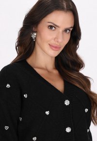 Schwarze Strickjacke mit V-Ausschnitt, strukturiertem Strick und verziert mit Herz- und Blumenförmigen Strasssteinen zur Dekoration.