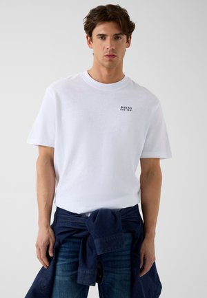 Jeune homme portant un t-shirt blanc Guess avec logo sur la poitrine, un jean foncé et une veste bleu marine nouée autour de la taille, debout devant un fond uni.