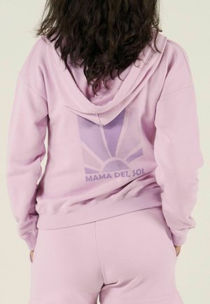 Vrouw met donker krullend haar, gekleed in een lichtpaarse hoodie en korte broek, met op de achterkant van de hoodie een zon en de tekst "MAMA DEL SOL".