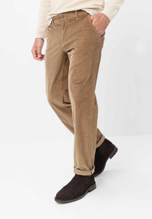 Mand iført beige kordfløjlsbukser og sorte ruskindsstøvler, med den ene hånd i lommen, stående foran en ensfarvet hvid baggrund.