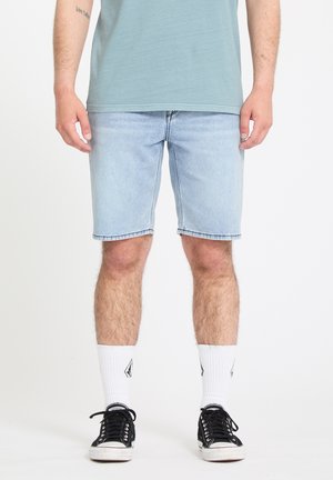 Homme portant un short en jean bleu clair, des chaussettes blanches montantes et des baskets noires, debout devant un fond blanc uni.