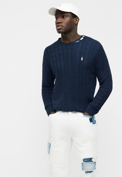 Άνδρας που φοράει μπλε σκούρο πλεκτό πουλόβερ, λευκό τζιν με ξεβάμματα και μπλε μπαλώματα, και λευκό καπέλο μπέιζμπολ, με τα χέρια στις τσέπες.