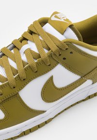 Ingrandimento di una sneaker Nike bianca e verde oliva con lacci texturizzati, logo swoosh cucito e etichetta in tessuto sulla linguetta.