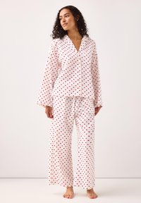 Hvid pyjamassæt bestående af en skjorte og brede bukser. Begge dele har et rødt hjerte-mønster og knapper foran. Blødt materiale med lange ærmer.