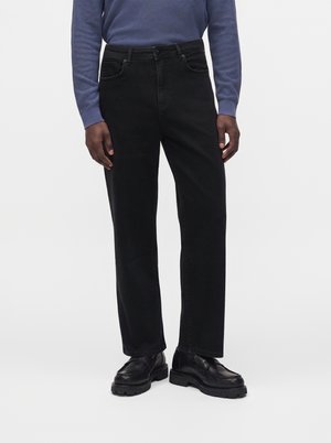 Homme portant un jean noir droit, des chaussures en cuir noires et une chemise bleue à manches longues, debout devant un fond blanc uni.