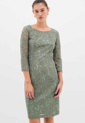 Vestito di pizzo in verde tenue, con maniche a tre quarti e una silhouette aderente. Presenta motivi floreali e vorticosi su tutto il capo.