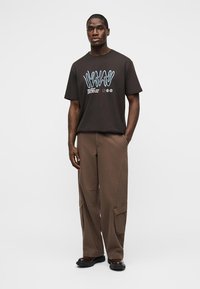 Άνδρας που φοράει μαύρο μπλουζάκι με σχέδιο και καφέ στρατιωτικό παντελόνι, στέκεται με το ένα χέρι στην τσέπη, μπροστά σε απλό φόντο.