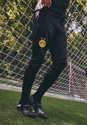 Schwarze Sporthosen mit einem gelben Borussia Dortmund Logo auf dem linken Oberschenkel, figurbetontes Design, kombiniert mit schwarzen Fußballschuhen und Socken.