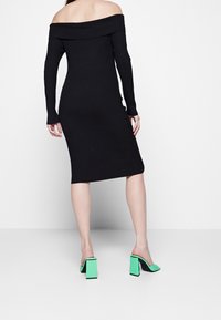 Černé žebrované šaty se střihy bez ramínek a dlouhými rukávy, s obepnutou siluetou, doplněné zelenými mules na blokovém podpatku.