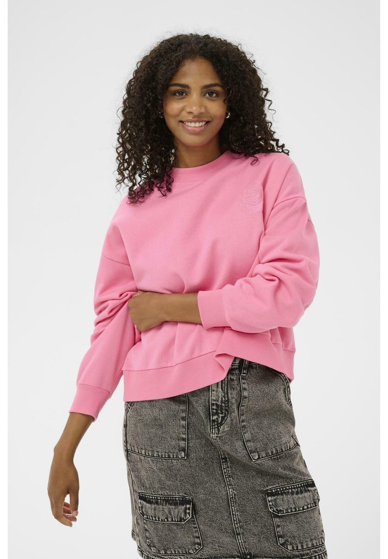 Sweat-shirt rose avec un col rond, des épaules tombantes et des poignets côtelés. Présentant un détail de rose brodé. Associé à une jupe grise.