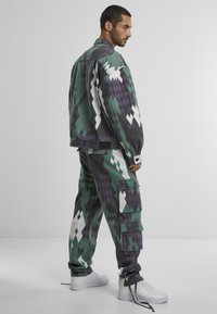 Chaqueta de múltiples patrones y pantalones cargo en verde, púrpura y blanco, con un ajuste relajado, botones a presión y bolsillos cargo. Zapatillas blancas.