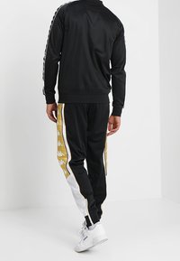 Black tracksuit with gold and white accents, featuring a zip-up jacket and tapered pants, made from smooth fabric with side stripes.