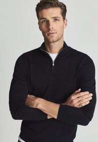 Homme aux cheveux courts et bruns, portant un pull noir à fermeture éclair et un maillot blanc, debout les bras croisés devant un fond clair uni.