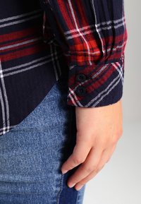 Geruite blouse met lange mouwen in rood, wit en marineblauw, met gebuttonde manchetten. Draagt blauwe spijkerbroek, met zichtbaar stofstructuur.