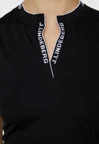 Black polo shirt featuring a half zip with a contrasting white trim. The collar includes brand text "J.LINDEBERG" in white. Soft fabric texture.