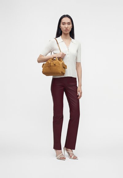 Femme portant un haut blanc à manches courtes, un pantalon bordeaux, des sandales blanches à talons hauts, tenant un sac à main en daim beige avec une bandoulière chaîne.