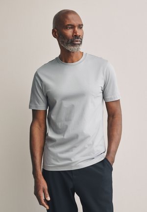Homme avec barbe grise portant un t-shirt gris clair à manches courtes et un pantalon noir, debout avec une main dans la poche devant un fond uni.