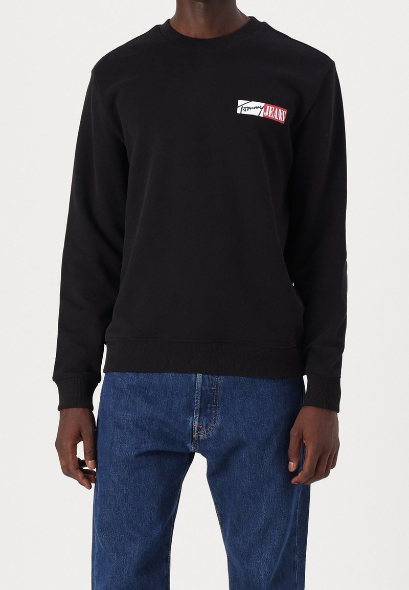 Sweat-shirt noir avec poignets et bord côtelés, arborant un logo coloré sur la poitrine gauche. Porté avec un jean en denim bleu.
