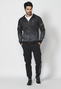 Čierna a šedá mikina na zips s gradientovým dizajnom, kapucňou na zaviazanie a rebrovanými manžetami. Kombinovaná s čiernymi cargo nohavicami a teniskami.