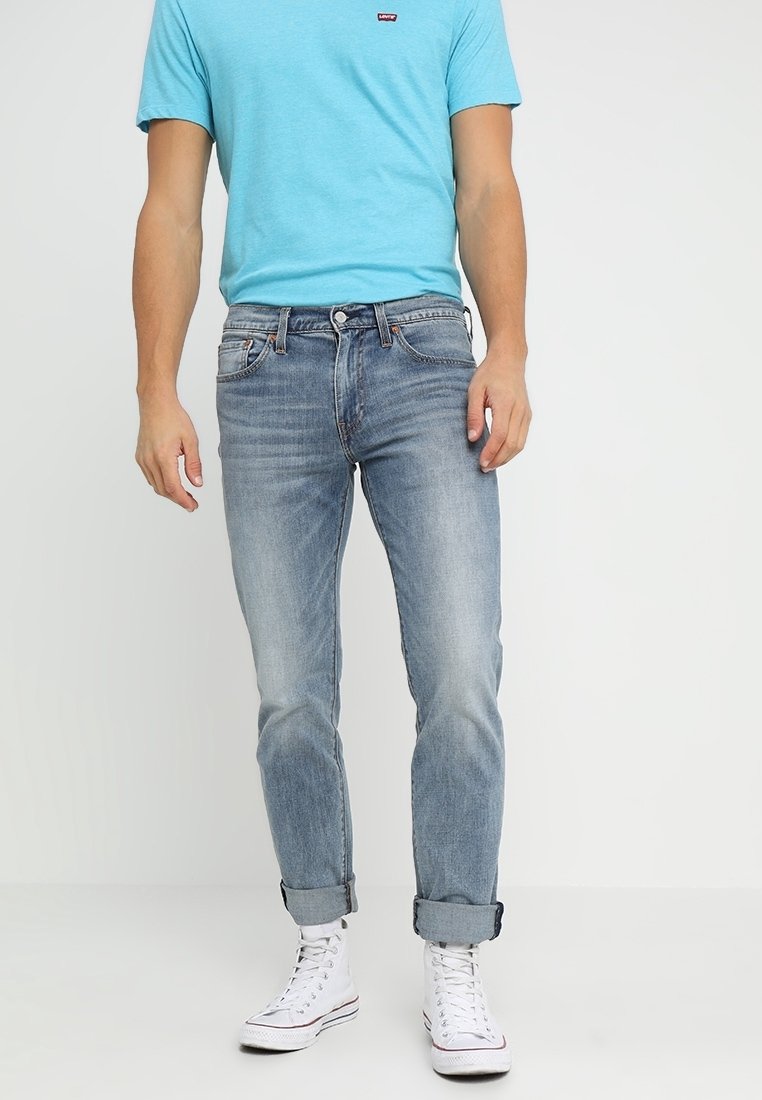 Homme portant un t-shirt bleu clair, un jean bleu délavé retroussé et des baskets blanches montantes sur fond uni.