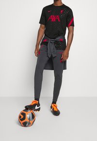 Černý dres Nike s červeným logem a akcenty, doplněný o šedé sportovní kalhoty. Fotbalové kopačky v černé a oranžové barvě. Tričko s tmavými vzorovanými detaily.