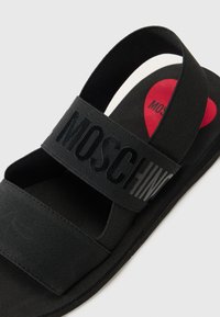 Mustat sandaalit, joissa on kaksi leveää hihnaa, joissa on kohokuvioitu "MOSCHINO"-teksti. Pohjassa on punainen sydän, jossa on musta "MOSCHINO"-teksti. Litteä muotoilu.