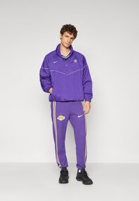 Lila Nike tréningruha, amely félcipzáras dzsekiből áll fehér díszítésekkel és logóval, valamint oldalsávos nadrággal, amelyen a Lakers logója szerepel.